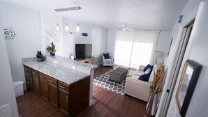 Una cocina moderna con encimera de granito, armarios de madera y horno integrado. Hay una zona de comedor con un sofá blanco y una mesa de centro. La habitación tiene un ventanal con cortinas de gasa y un ventilador de techo.