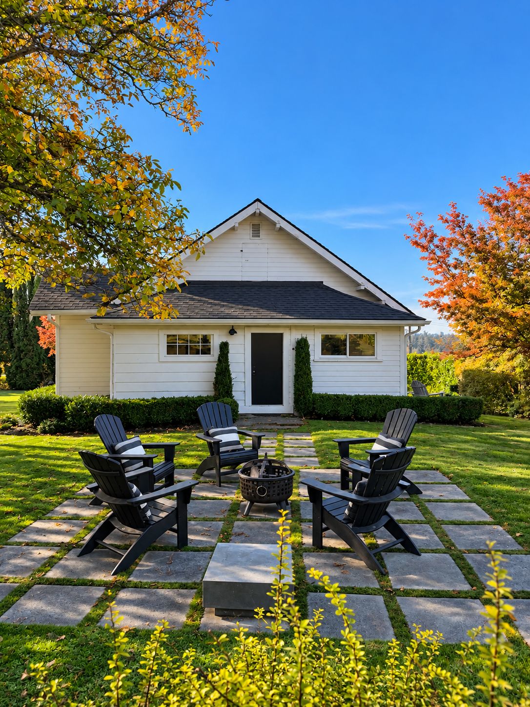 Une maison blanche avec un porche à l’avant, quatre chaises Adirondack, un foyer et un chemin de pierres.