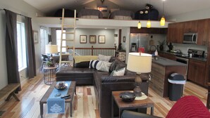 A cozy living room with a sofa, a coffee table, and a dining table. There is a kitchen area with a refrigerator and a microwave. A ladder is visible in the room, leading to a loft bed. - Custom home in St Mary’s Glacier (Idaho Springs)