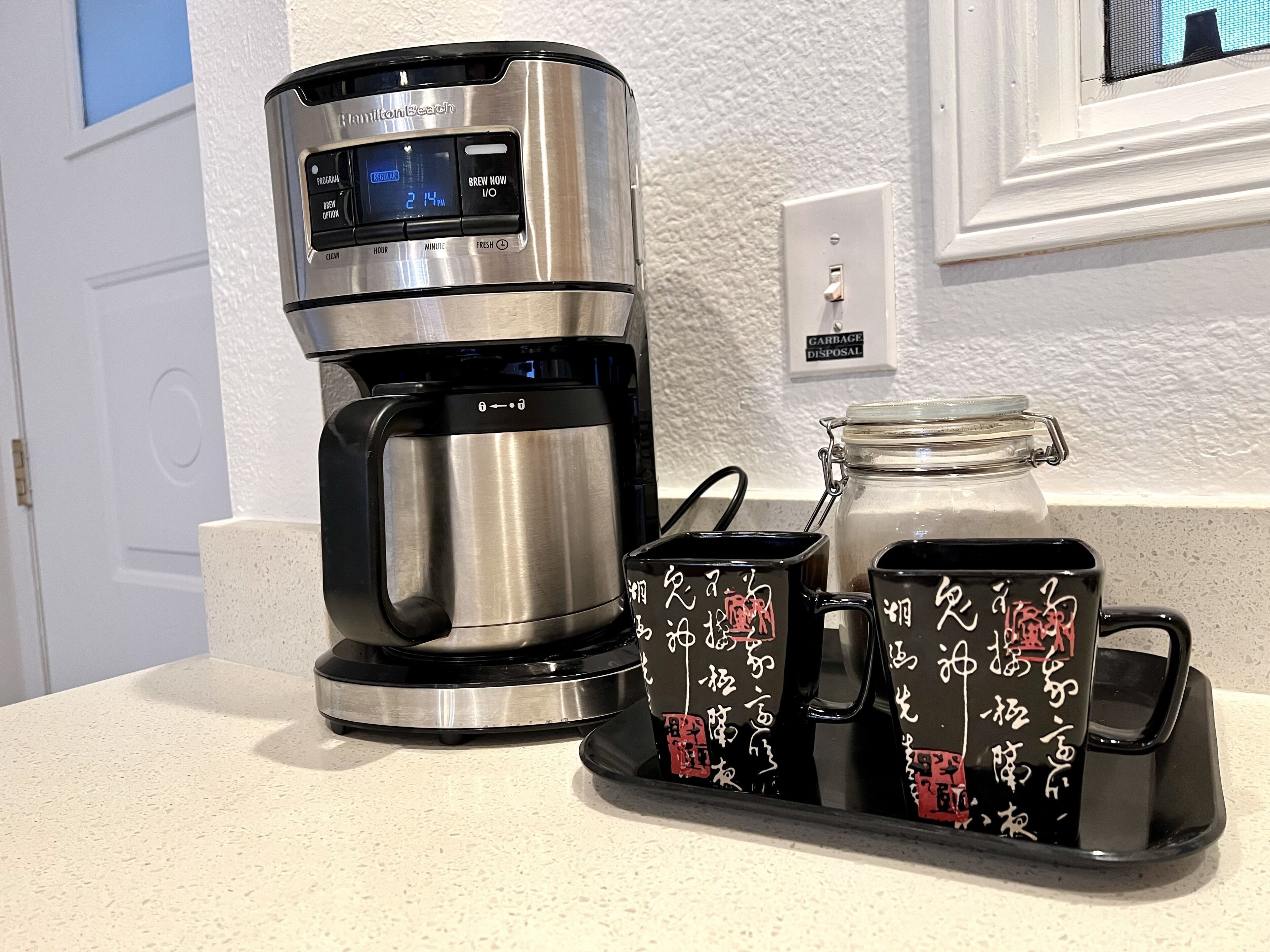Una macchina da caffè Hamilton Beach con display digitale, un cestino e due tazze con caratteri asiatici.