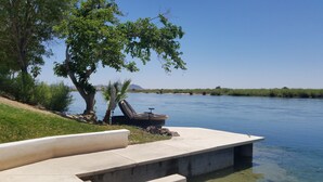 A riverside patio with a concrete bench, a lounge chair, and a small table. - River Front guest house with Ramp, Beach, Volleyball, and Gazebo on the Water (Big River)