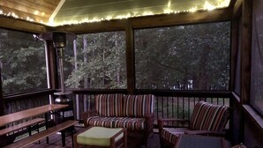 A covered outdoor seating area with wooden benches and wicker furniture, featuring large windows and string lights.