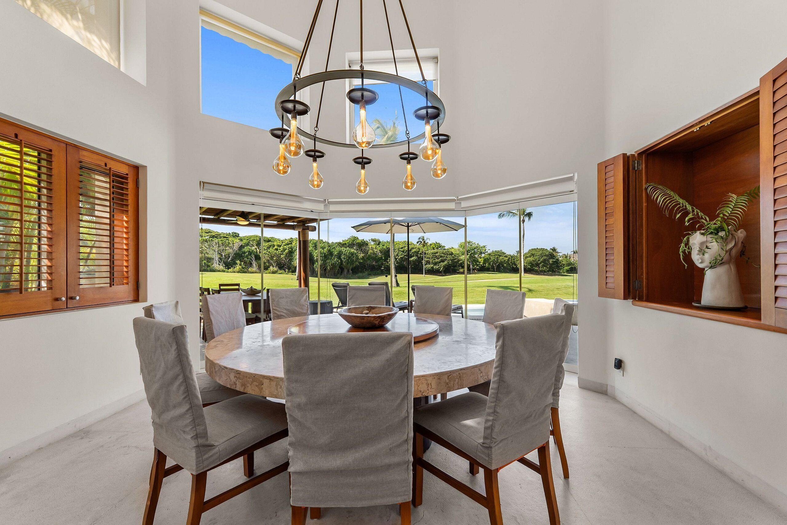 Een ronde eettafel met stoelen, een kroonluchter en uitzicht op een golfbaan door grote ramen.