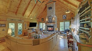A log cabin interior with a stone fireplace, wooden ceiling beams, and a cozy seating area.