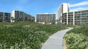 Un chemin mène à un complexe hôtelier moderne, doté de plusieurs étages et de balcons, et entouré de verdure et de palmiers.