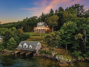 Une grande maison à étages, dotée d’une tour imposante, entourée d’une forêt dense et bordée d’un quai qui s’avance sur une étendue d’eau.