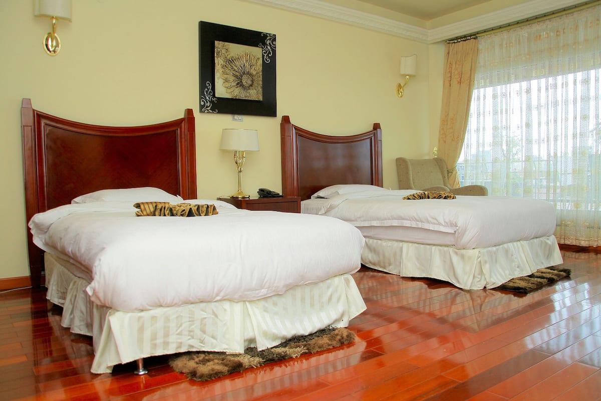Habitación de hotel con dos camas, cabecera de madera, mesita de noche con lámpara y un ventanal con cortinas.