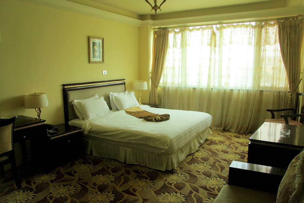 Habitación de hotel con una cama grande, una mesita de noche, una lámpara y una ventana con cortinas.