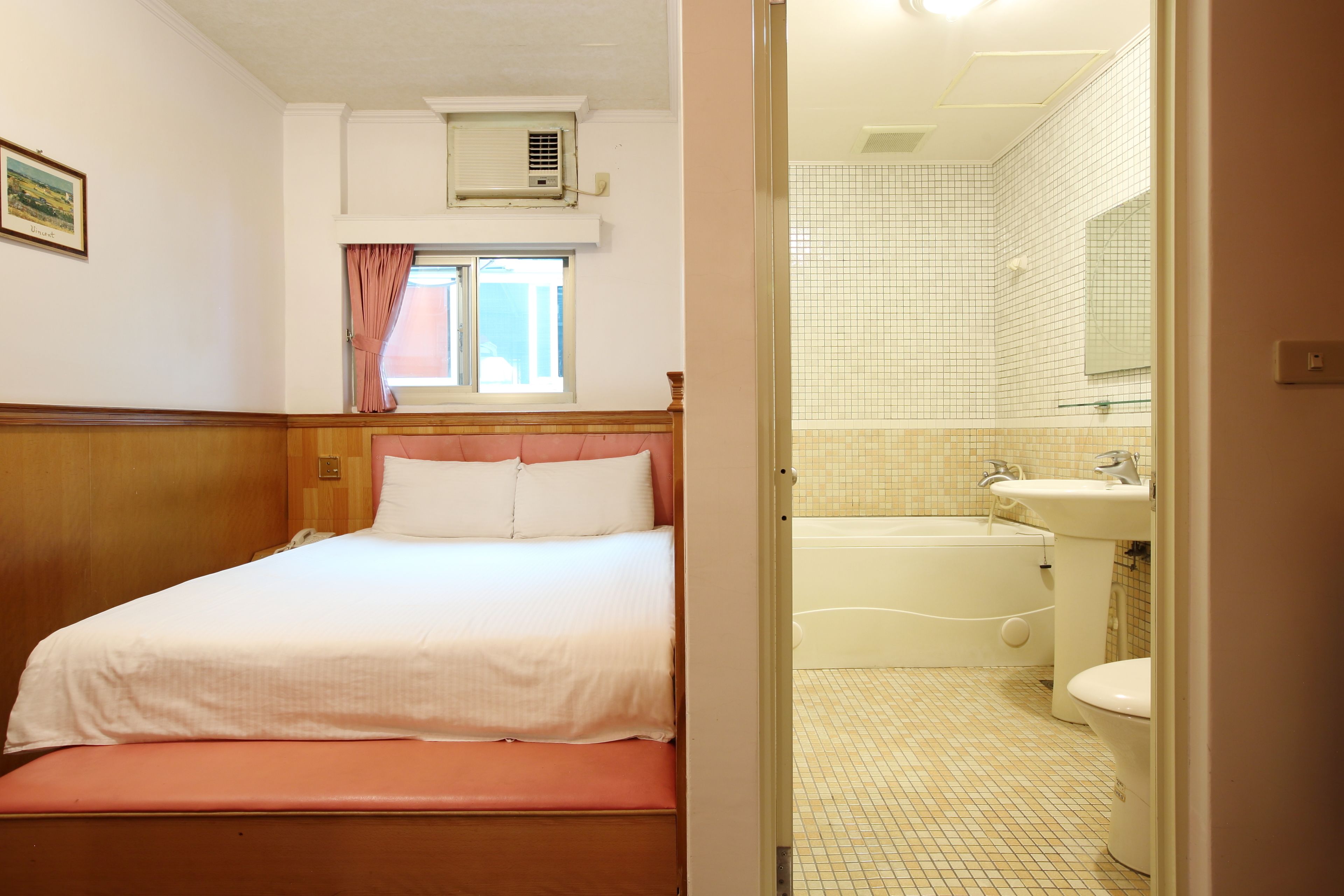 Una habitación de hotel con cama, ventana con cortinas, unidad de aire acondicionado, baño con bañera, lavamanos e inodoro.