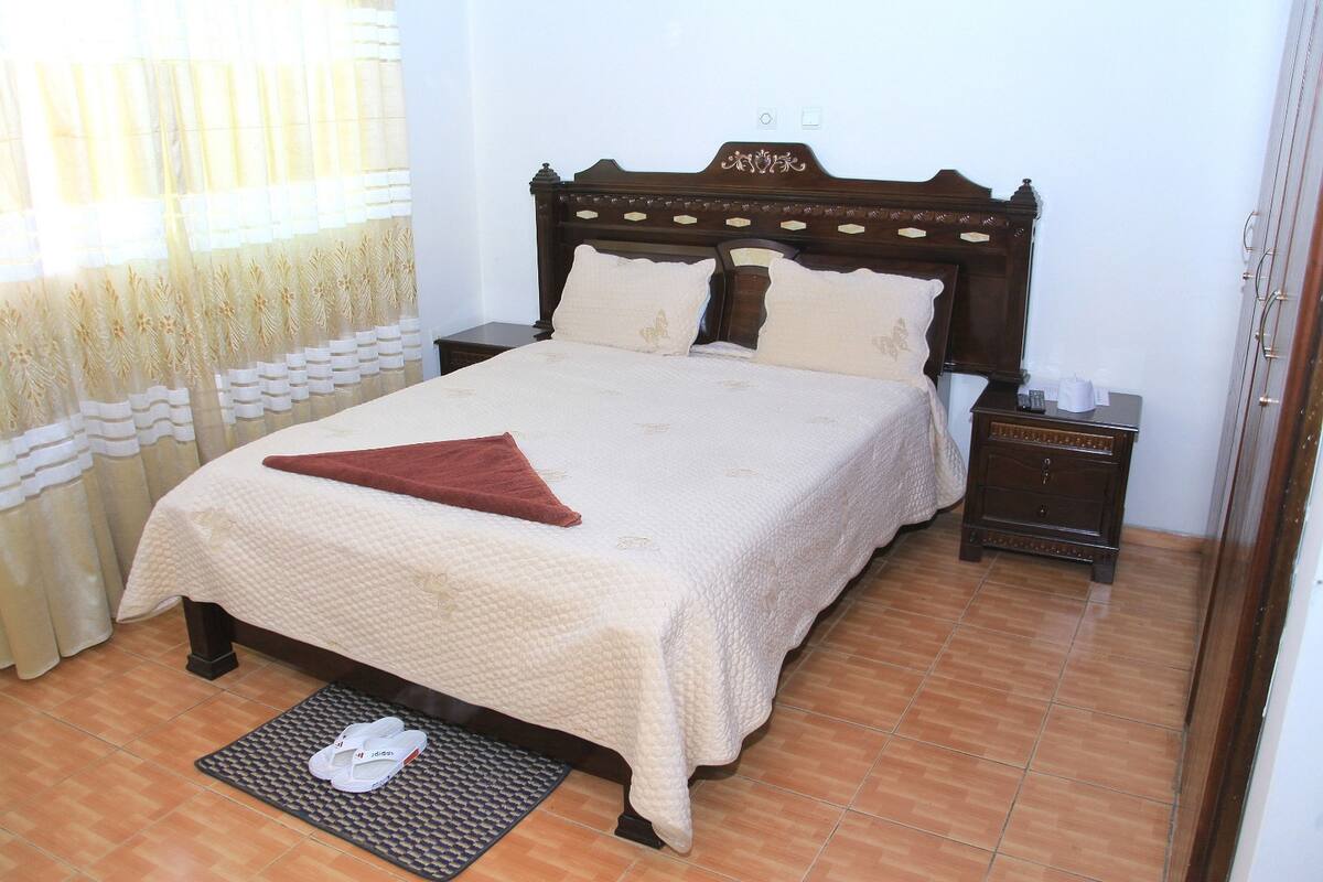 A neatly made bed with a brown headboard, a bedside table with a lamp, and a pair of slippers on a mat.