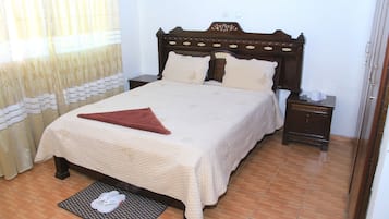 A neatly made bed with a brown headboard, a bedside table with a lamp, and a pair of slippers on a mat.