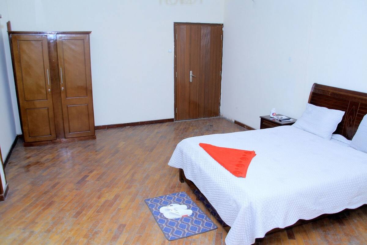 A bedroom with a wooden wardrobe, a single bed with a white and orange blanket, and a small bedside table with a phone and a tissue box.