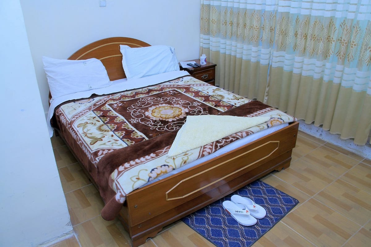 A neatly made bed with a patterned bedspread, a wooden headboard, and a pair of slippers on a blue mat.
