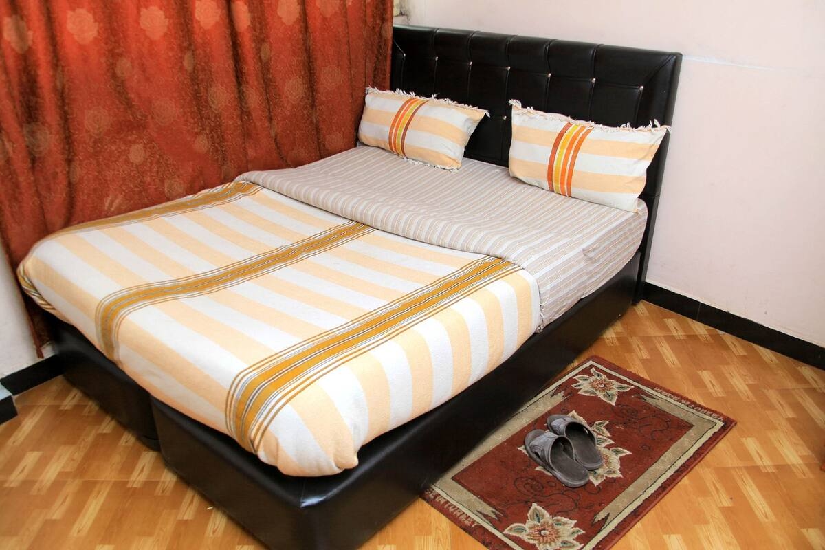 A neatly made bed with striped bedding and two pillows, a black upholstered headboard, and a wooden floor with a rug.