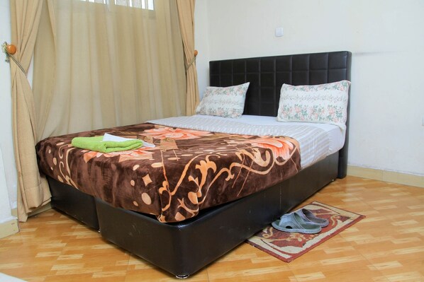 A bed with a patterned bedspread, a pair of slippers on a rug, and curtains on the window.