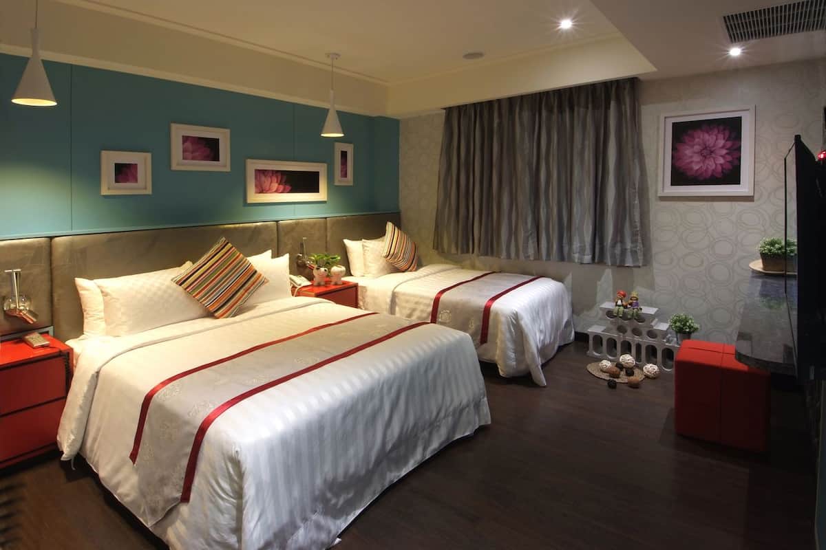 Une chambre d’hôtel avec deux lits, une table de chevet rouge, un pouf rouge et une grande fenêtre avec des rideaux.