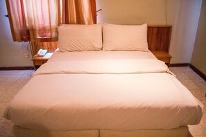 A neatly made bed with a wooden headboard, a bedside table with a phone, and a curtain partially drawn to the side. - Chelsea Suites Limited (Lagos)