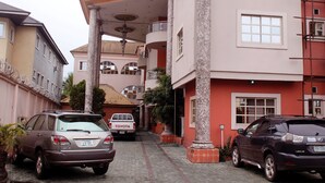 A multi-story building with a central courtyard, a Toyota truck parked in front, and a Lexus SUV parked on the side. - Paloma Hotels (Port Harcourt)
