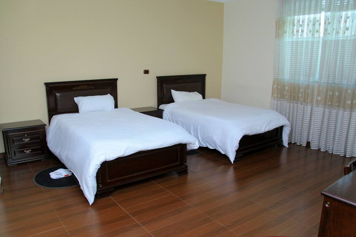 A hotel room with two single beds, wooden headboards, a small bedside table with drawers, a round rug, and a window with sheer curtains.