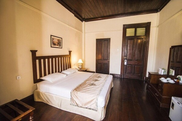 A bedroom with a wooden bed, a nightstand with a lamp, a wooden wardrobe, and a door. - Queens Hotel (Kandy)