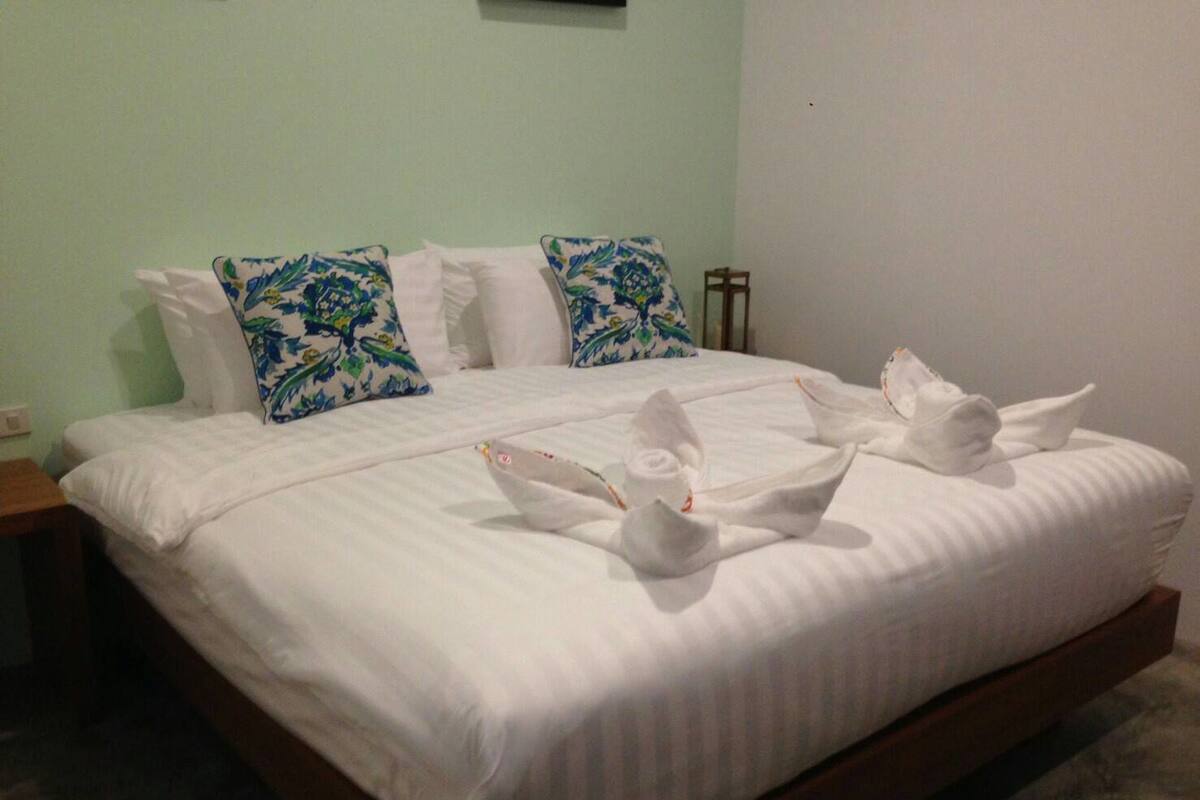 A neatly made bed with white linens and floral-patterned pillows, a wooden nightstand, and a small table with a lamp.