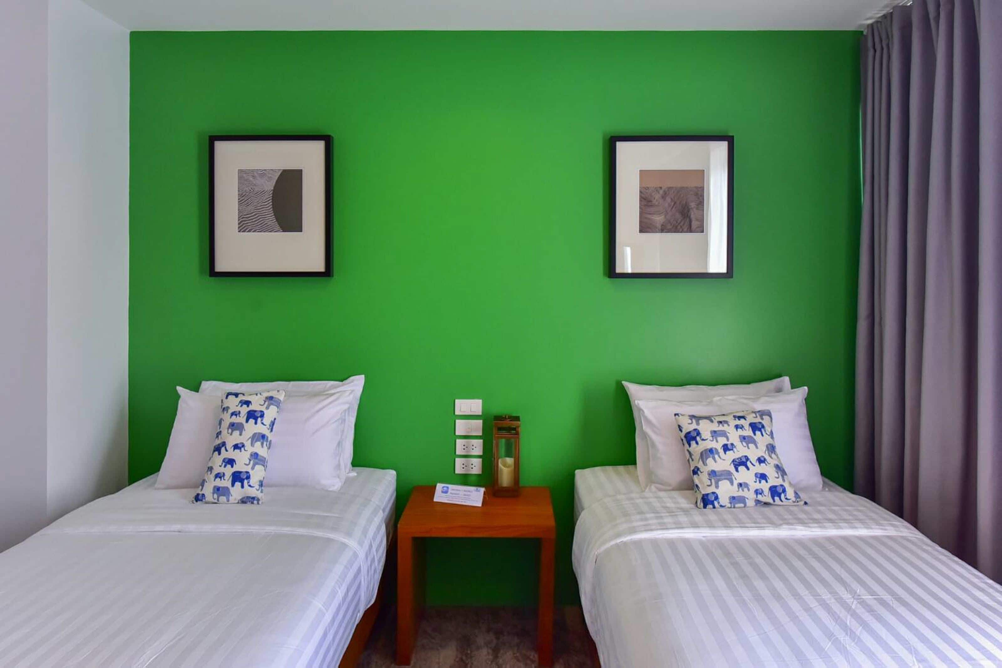 A room with two beds, green walls, framed pictures, and a small table with a lamp.
