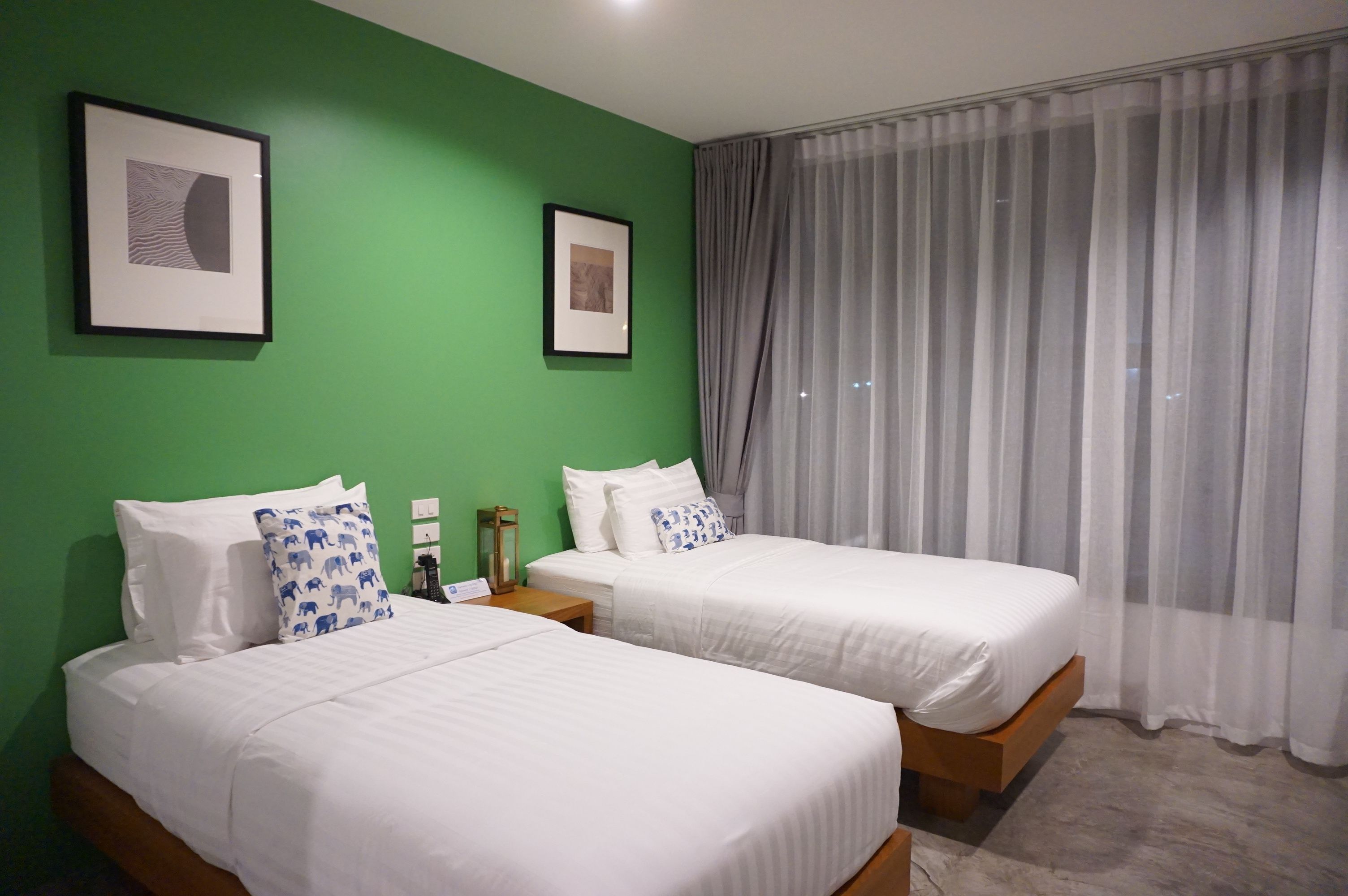 A hotel room with two beds, green walls, framed artwork, and a large window with sheer curtains.