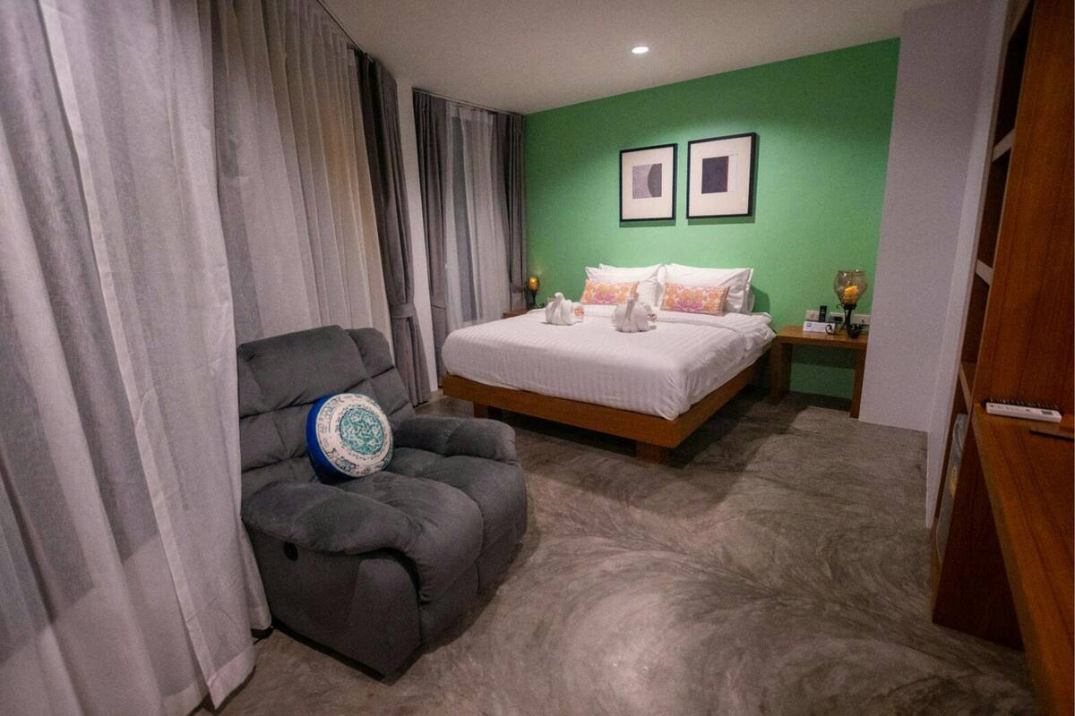 A hotel room with a bed, a grey armchair, a nightstand, and a green wall.