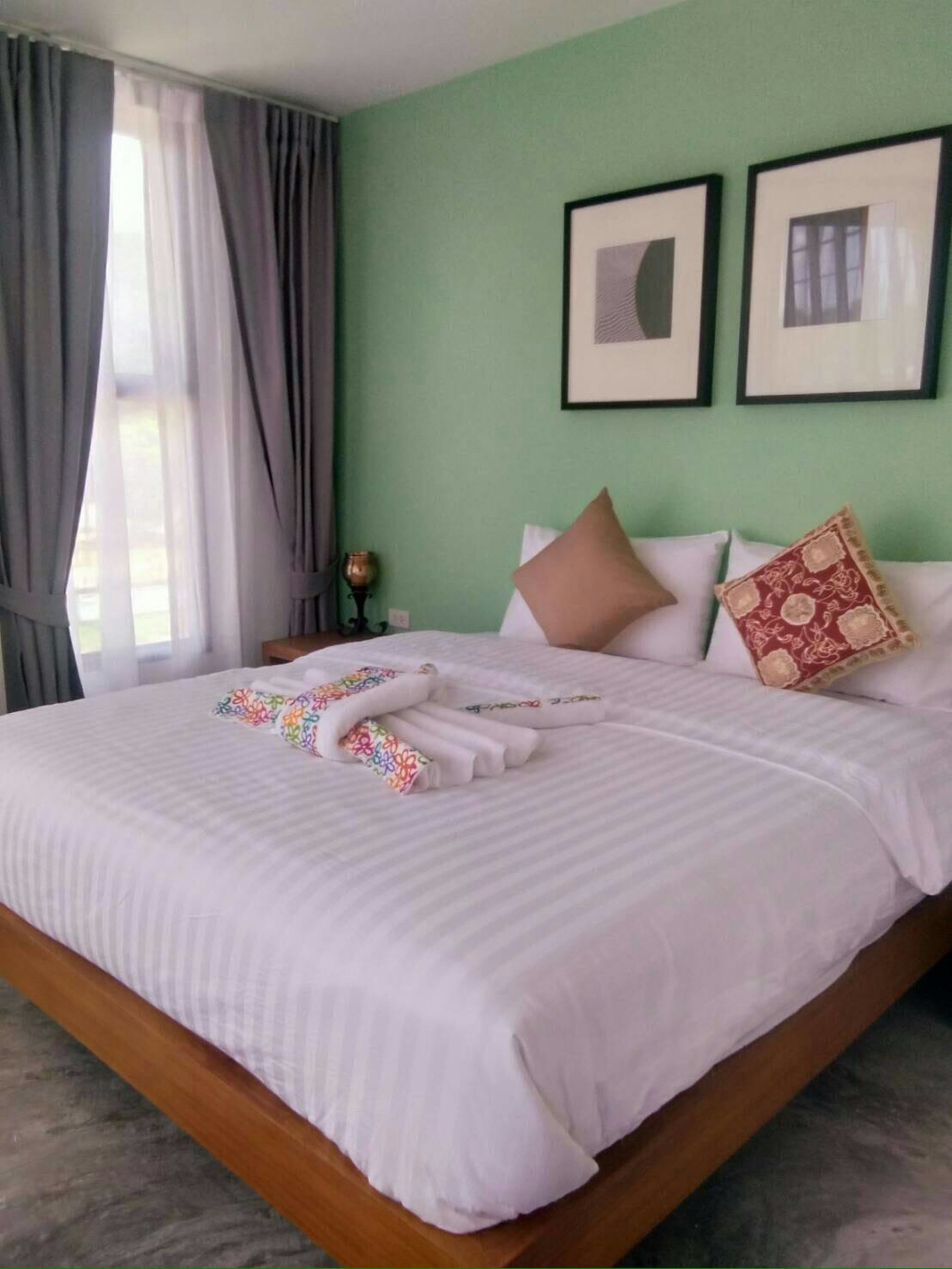 Uma cama bem arrumada com roupa de cama branca e uma toalha colorida, uma mesa de cabeceira com abajur e duas fotografias emolduradas na parede.