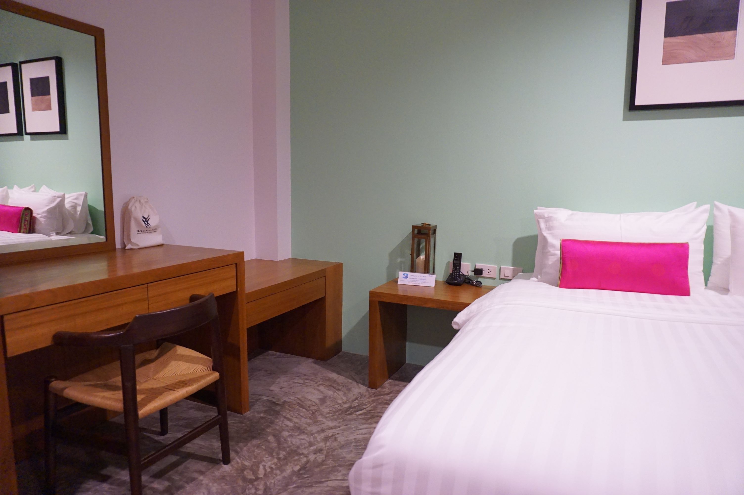 A hotel room with a bed, a desk, a chair, a bedside table, a mirror, and a wall with framed pictures.