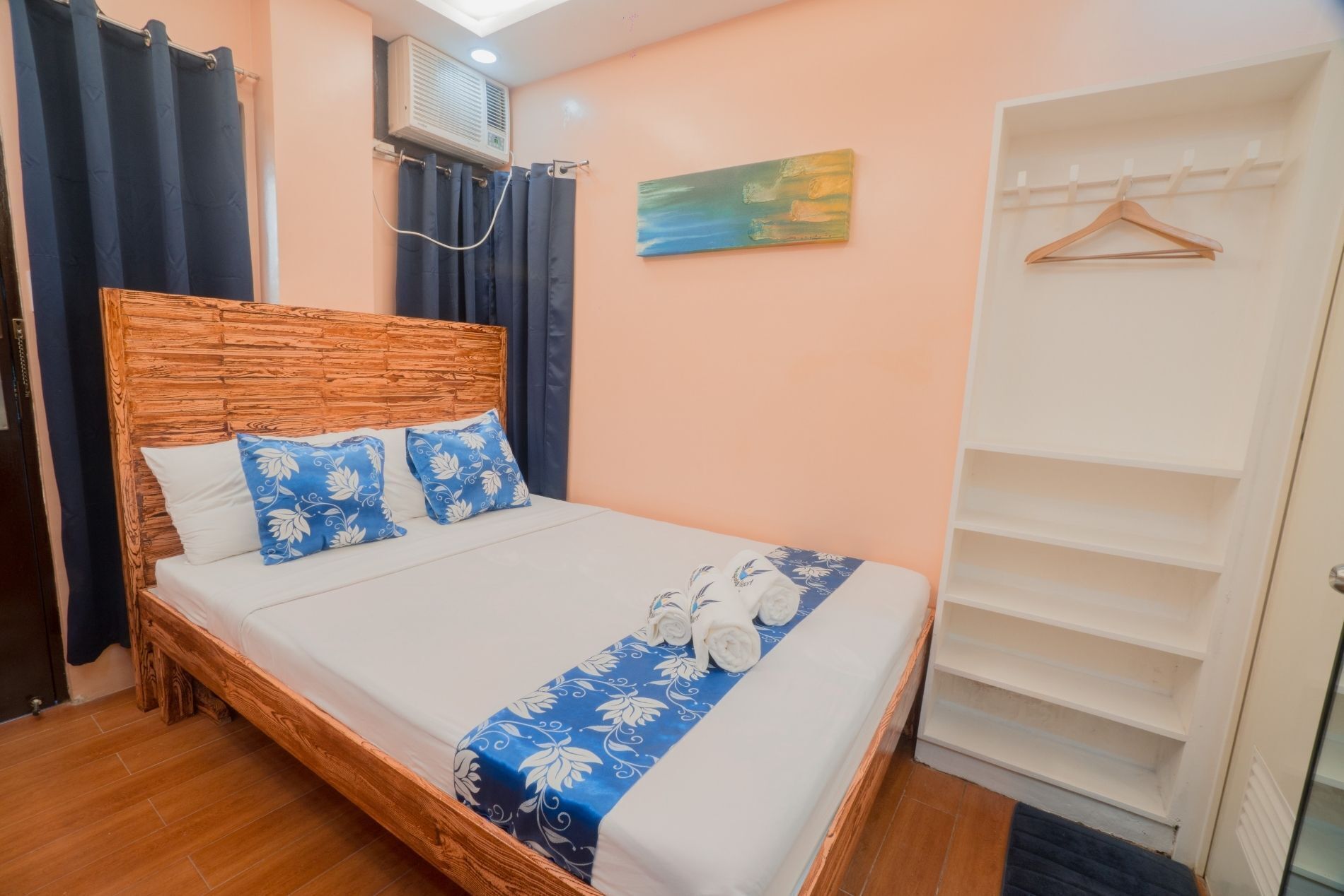 Une chambre avec une tête de lit en bois, un lit agrémenté d’oreillers à motifs bleus et blancs, une couvre-lit bleue et blanche, un climatiseur fixé au mur, une porte avec une armoire blanche et un tableau accroché au mur.
