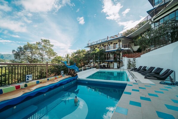 Una piscina con una escultura de delfín, un letrero de 60 centímetros de altura, y un edificio con un balcón que da a la piscina.