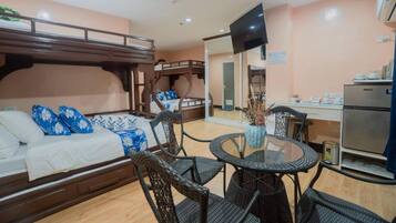 A hotel room with bunk beds, a dining area with a round table and chairs, a kitchen area with a refrigerator, and a flat-screen TV mounted on the wall.