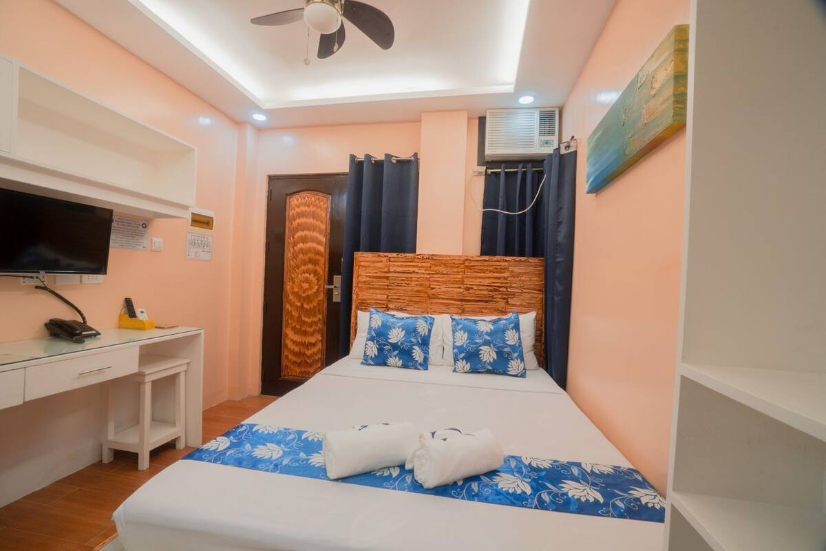 A hotel room with a bed, a desk, a television, and an air conditioning unit.