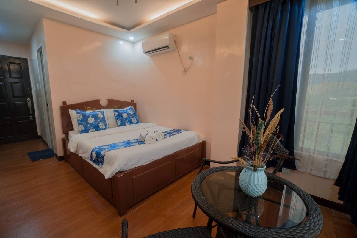 A hotel room with a wooden bed, a small round table with a vase, and a window with curtains.