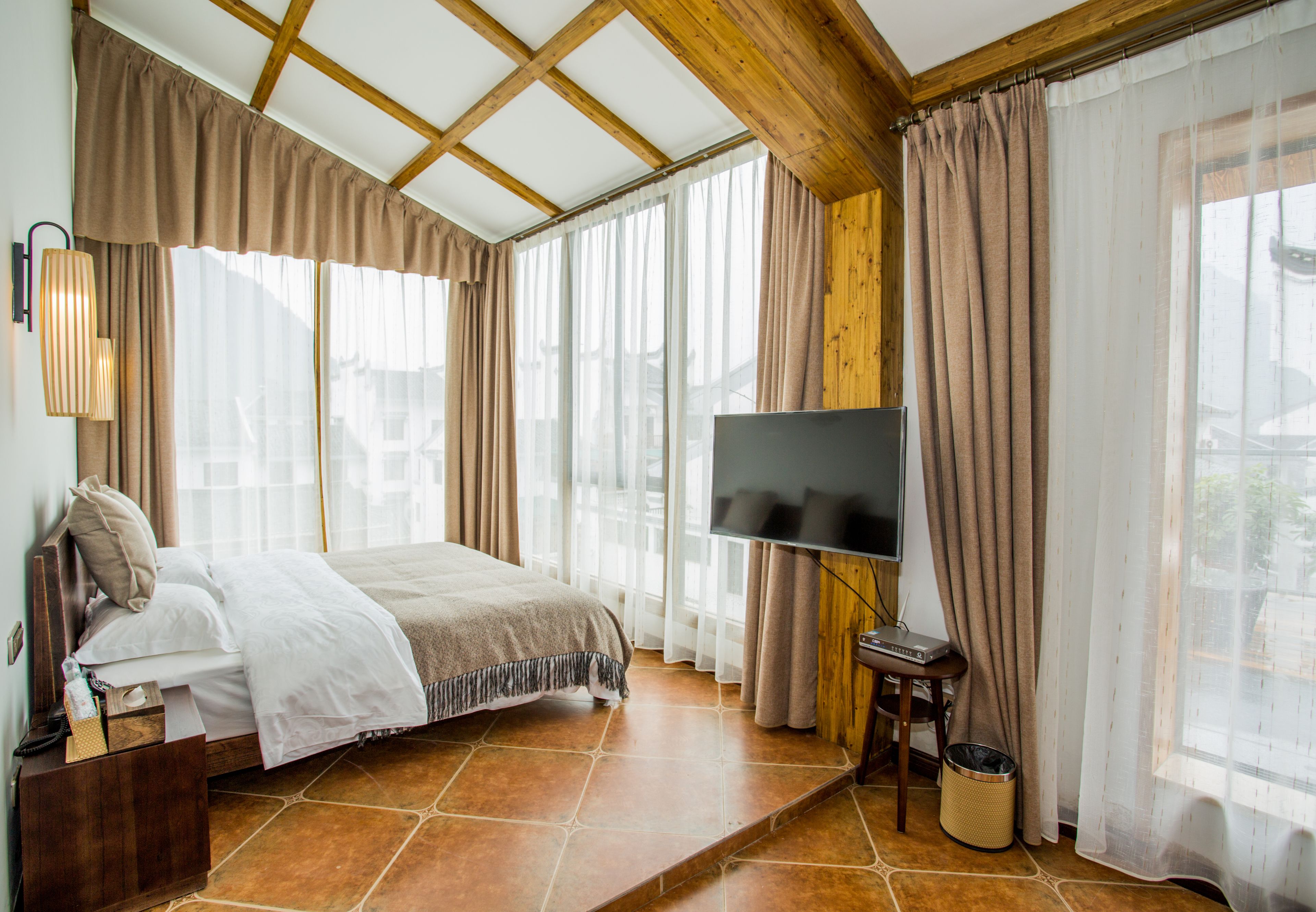 A bedroom with a bed, a TV, a wooden ceiling, and large windows with curtains.