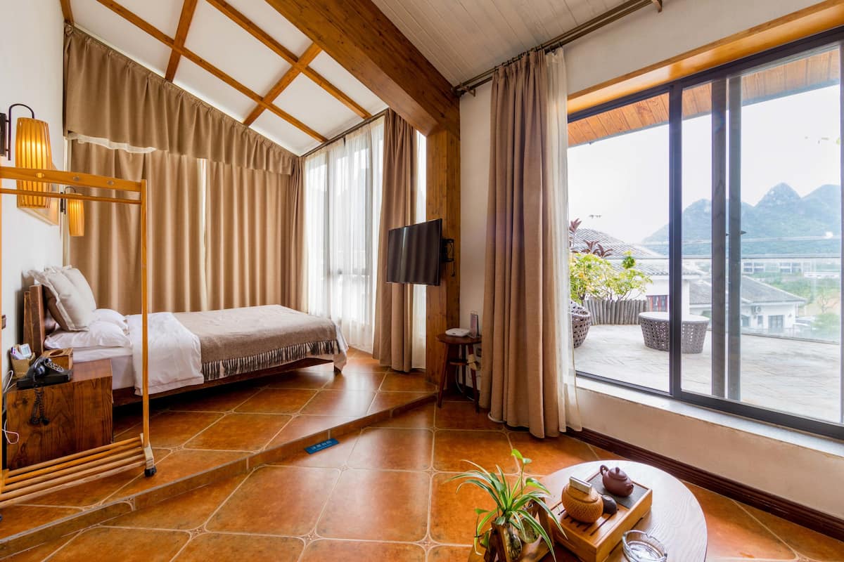 A hotel room with a bed, a TV, a wooden desk, a plant, and a view of mountains through large windows.