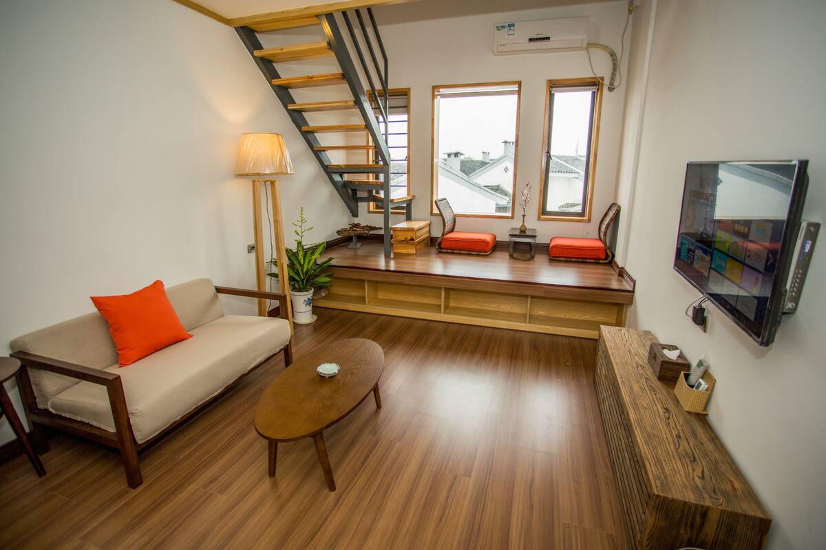 A modern living room with a sofa, coffee table, and a wooden bench. A staircase leads to an upper level, and there is a TV mounted on the wall.
