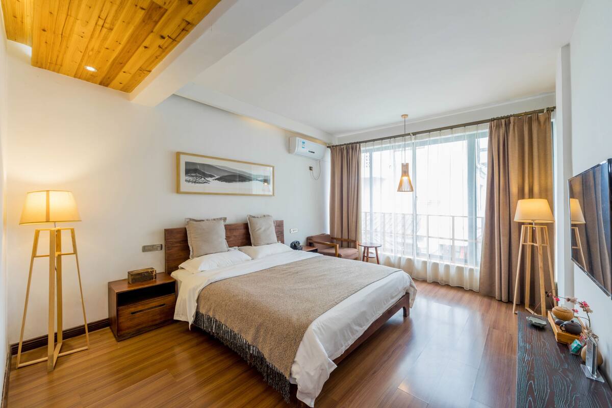 A hotel room with a wooden ceiling, a large bed with a beige blanket, a bedside table with a lamp, a chair, and a window with curtains.