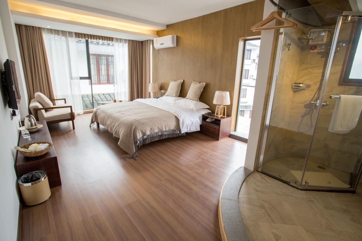 A hotel room with a large bed, a sitting area with a chair and a table, a glass-enclosed shower, and a view of the outside through large windows.