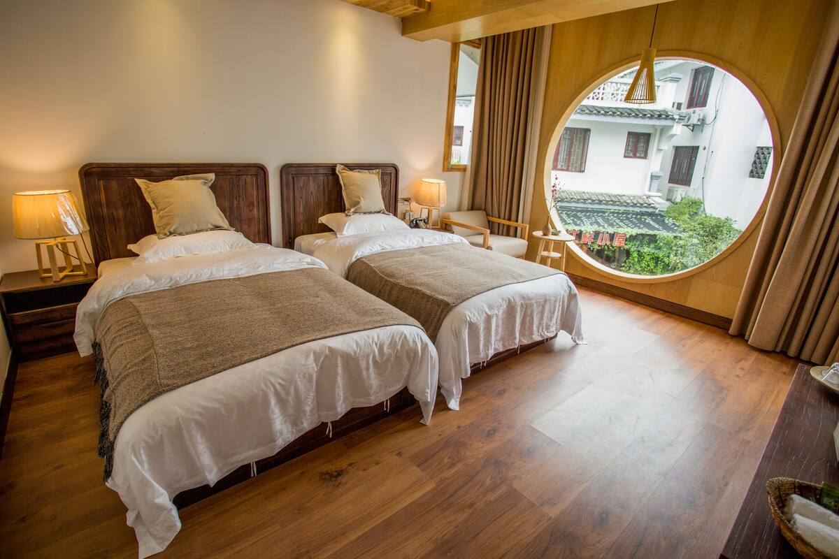 A hotel room with two beds, wooden flooring, a round window with a view of buildings, and a small table with a lamp.