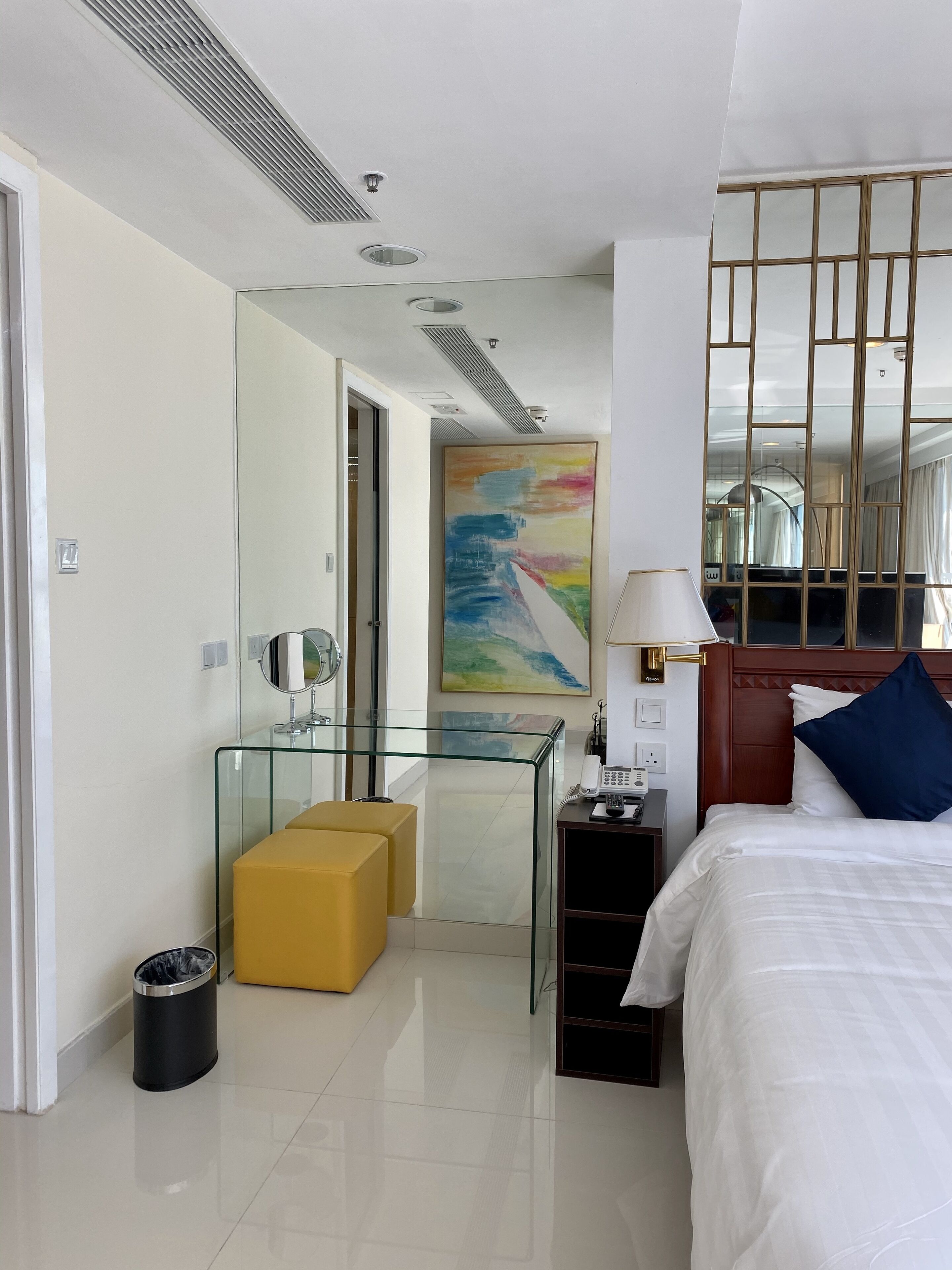A modern hotel room with a bed, a glass partition, a small yellow ottoman, a desk with a lamp, and a painting on the wall.
