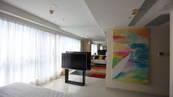Habitació d'hotel moderna amb un llit gran, televisió de pantalla plana, un quadre abstracte i vistós, i vista de la ciutat a través de cortines translúcides.