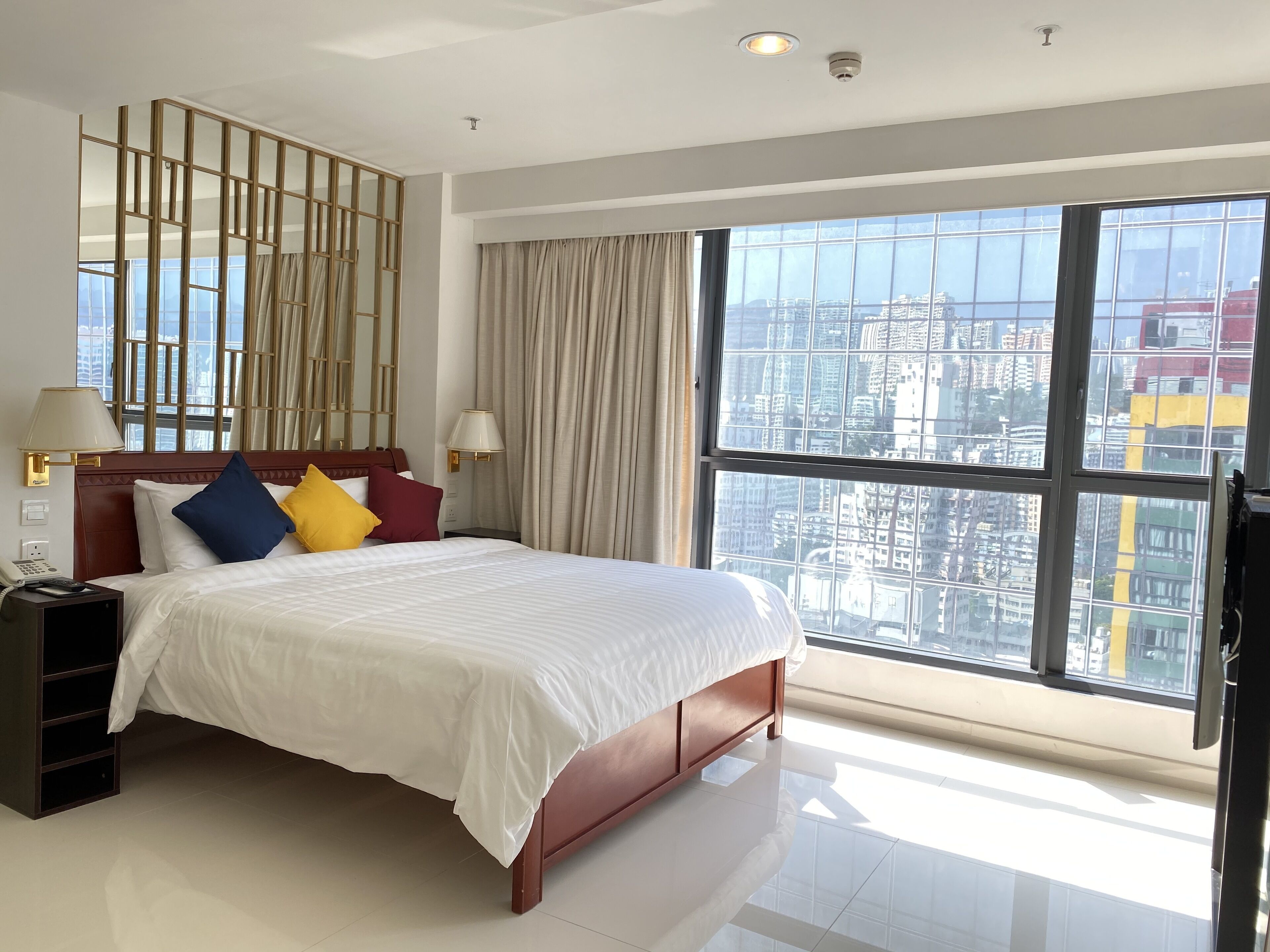 A hotel room with a large bed, a nightstand, a lamp, and a view of the city through large windows.
