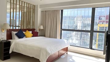 A hotel room with a large bed, a nightstand, a lamp, and a view of the city through large windows.