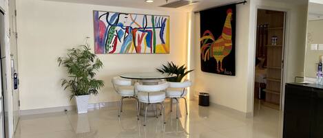 Eine moderne Hotelhalle mit einem runden Esstisch, weißen Stühlen, einer Küstertopf-Pflanze, einem farbenfrohen abstrakten Gemälde und einer Kunst-Darstellung eines Hahns.