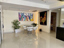 A modern hotel lobby with a round dining table, white chairs, a potted plant, a colorful abstract painting, and a rooster artwork.