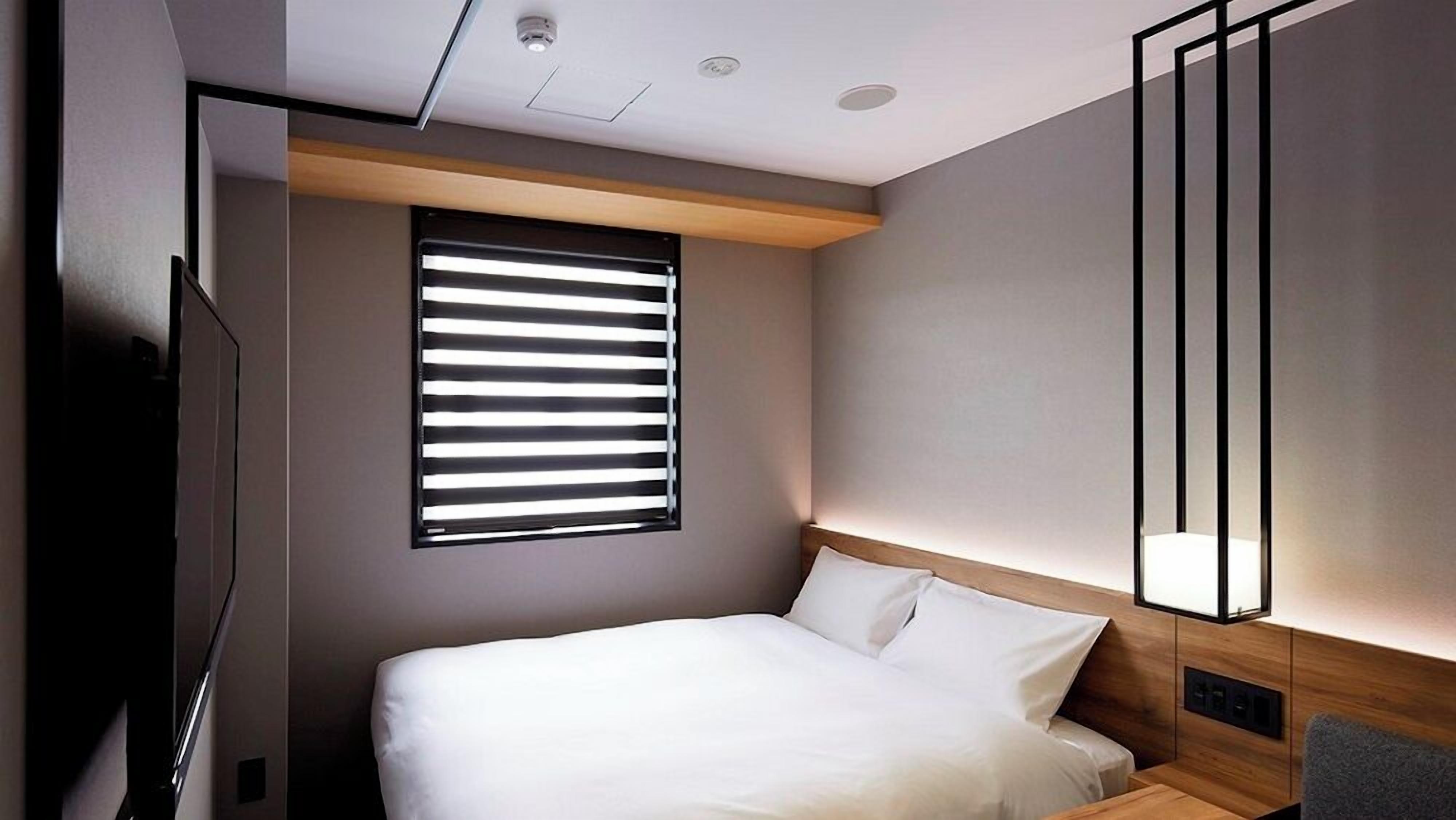A modern hotel room with a bed, a window with blinds, a bedside table, and a wall-mounted light fixture.
