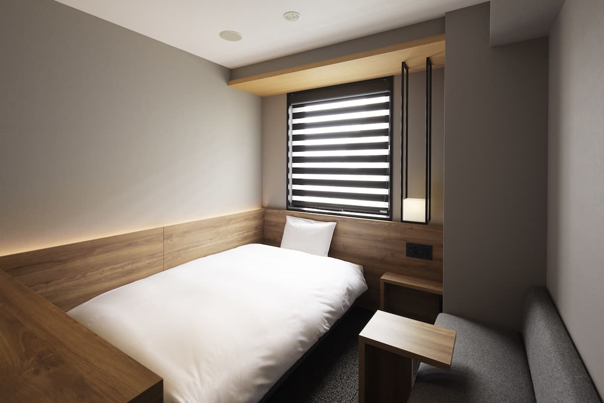 A modern bedroom with a wooden headboard, a single bed, a bedside table, and a window with blinds.