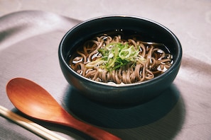 Un tazón de sopa de fideos oscura con ingredientes verdes, acompañado de una cuchara de madera y palillos sobre una mesa.